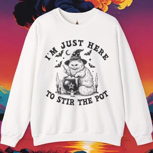 May include: White sweatshirt with a graphic of a cat wearing a witch hat, stirring a cauldron. The text reads "I'm just here to stir the pot." Bats, candles, and a crescent moon are also featured in the design.