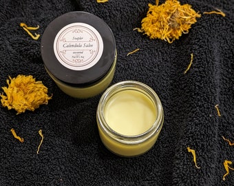 以下が含まれることがあります： 黒い蓋が付いた、無香料の calendula 軟膏が入った小さなガラス瓶。瓶には「Soughter Calendula Salve unscented Net Wt. 4 oz.」とラベルが貼られています。
