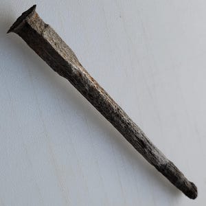 Puede incluir: Primer plano de un clavo antiguo y desgastado. El clavo tiene una cabeza rectangular y un vástago largo y cónico. El metal es de un gris oscuro envejecido con textura visible y signos de óxido, lo que sugiere edad y uso.