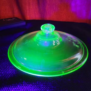 Könnte beinhalten: Ein leuchtend grüner Glasdeckel mit rundem Oberteil und kleinem Knauf. Der Deckel ist kreisförmig und scheint aus dickem Glas zu bestehen. Das Objekt befindet sich auf einer dunklen Oberfläche mit rotem und violettem Hintergrund.