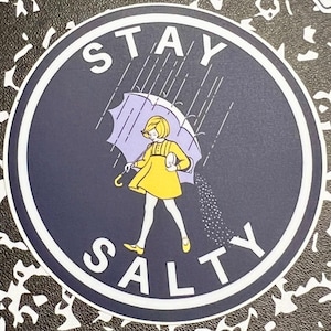 May include: A round sticker with a blue background and white text that says "STAY SALTY". A girl in a yellow dress is holding an umbrella and walking away from a rain cloud. The rain is falling as salt.