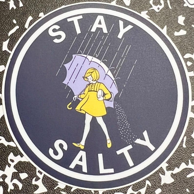 May include: A round sticker with a blue background and white text that says "STAY SALTY". A girl in a yellow dress is holding an umbrella and walking away from a rain cloud. The rain is falling as salt.