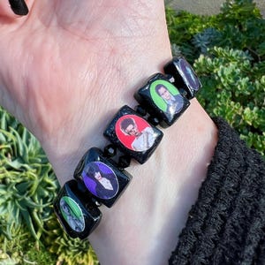 Op de afbeelding: Zwart kralenarmband met rechthoekige zwarte kralen met kleurrijke afbeeldingen van personages. De armband wordt gedragen om een pols met een zwarte mouw. Groene planten vormen de achtergrond.
