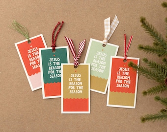 Op de afbeelding: Vijf rechthoekige kerstcadeaulabels met de tekst "JESUS IS THE REASON FOR THE SEASON". Elk label heeft een ander kleurenschema, waaronder rood, groen en goud, met decoratieve linten.