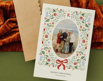 May include: A Christmas card with a family photo inside an oval frame, surrounded by a floral wreath with birds and a red bow. The card is on a green surface with a brown envelope and a rust-colored velvet cloth.