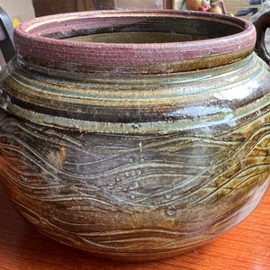 May include: A large, round, brown ceramic pot with a wide opening and a wavy, textured design. The pot has a dark brown glaze with streaks of green and white.