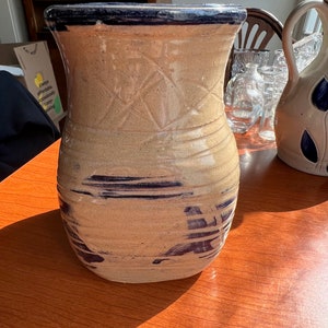 May include: A brown ceramic pitcher with a blue glaze and a dark blue rim. The pitcher has a textured surface and a wide base.