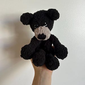 May include: A black crocheted stuffed bear with a gray nose and black eyes. The bear is sitting up with its arms outstretched.