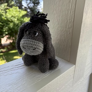 May include: A handmade crochet donkey doll in shades of gray. The donkey has a gray muzzle, dark gray body, and black yarn hair. The doll is sitting on a white surface with a blurred green background.