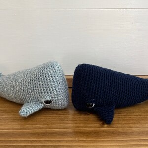 May include: Two crocheted whale plushies, one grey and one navy blue, with black eyes. The whales are lying on a wooden surface.