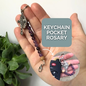 May include: A purple and brown beaded keychain rosary with a silver cross charm. The rosary is attached to a silver key ring. KEYCHAIN POCKET ROSARY is written in white text on a blue background.