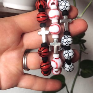 Puede incluir: Tres pulseras con dijes de cruz plateados y cuentas con temática deportiva. Las cuentas son rojas, blancas y negras y presentan diseños de baloncesto, béisbol y fútbol.