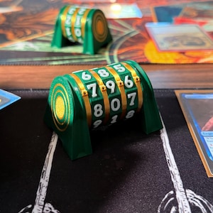 Op de afbeelding: Groene en gouden dobbelsteenroller met witte cijfers. De dobbelsteenroller heeft een cilindrisch ontwerp met een reeks draaiende nummerwielen. De dobbelsteenroller is ontworpen voor tabletop gaming.
