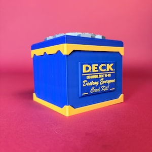 May include: A blue and yellow rectangular storage box with a lid. The front features a yellow plaque with the text D.E.C.K. THE ORIGINAL (VER 1.76-CC) Destroy Everyone Card Kit! The box has a smooth finish and a distinct yellow trim along the top and bottom edges.