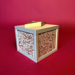 May include: A cube-shaped box featuring light gray smooth edges and recessed side panels with a complex, intricate copper-colored labyrinthine pattern. A small, angled, metallic gold-toned rectangular piece sits on the top surface of the cube.