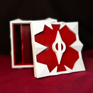 May include: White box with a red interior and a white geometric design on the lid. The design features a white star shape with a red center and a white arrow in the center.