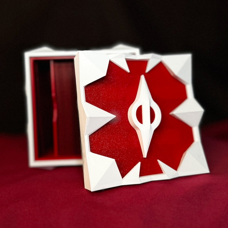 May include: White box with a red interior and a white geometric design on the lid. The design features a white star shape with a red center and a white arrow in the center.