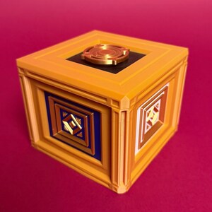 May include: A cubic box featuring a bright orange frame with a circular gold-colored emblem on the top. One side displays a blue square panel with a gold geometric design, while the adjacent side features a white panel with a similar gold pattern.