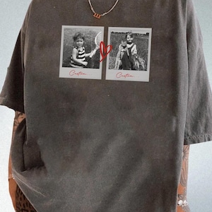 May include: Dark gray t-shirt featuring two black and white Polaroid-style photos connected by a red heart graphic. The photos depict children. The word "Custom..." is printed below each photo. The shirt has short sleeves and a relaxed fit.
