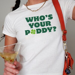 May include: White t-shirt with the text "WHO'S YOUR PADDY?" in green, featuring a shamrock. The shirt is paired with a red purse and a gold chain necklace. A martini glass with olives is held in the hand.