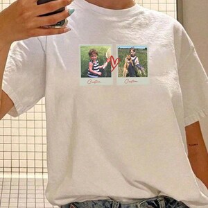 May include: White t-shirt featuring two polaroid-style photos connected by a red heart. The photos show children in outdoor settings. The word "Custom..." is printed below each photo. The shirt is made of a soft, comfortable material.