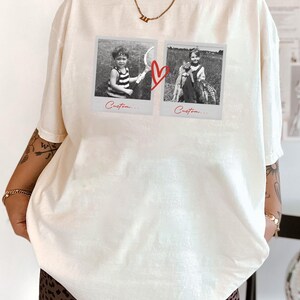 May include: Cream-colored t-shirt featuring two black and white polaroid-style images connected by a red heart graphic. The images show children. The word "Custom..." is written below each image. The shirt is a relaxed fit.