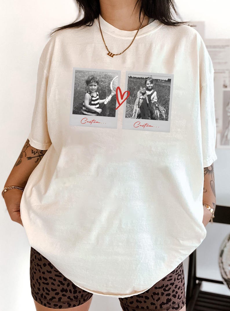 May include: Cream-colored t-shirt featuring two black and white polaroid-style images connected by a red heart graphic. The images show children. The word "Custom..." is written below each image. The shirt is a relaxed fit.