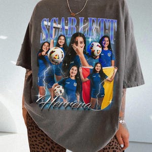 Könnte beinhalten: Dunkelgraues T-Shirt mit einem Grafikdesign im Vintage-Stil. Die Grafik zeigt den Namen "SCARLETT" in Blau und Weiß, darunter "Herrera". Das Design enthält Bilder einer Fußballspielerin in blauem Trikot mit einem Fußball und einer Flagge.