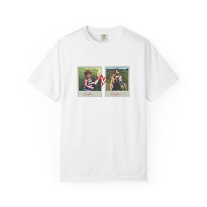 May include: White t-shirt featuring two framed photos with children and a red heart graphic. The photos show children in a field. The word "Cristina" is written below each photo.