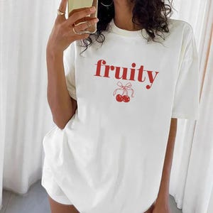 Op de afbeelding: Wit T-shirt met het woord "fruity" in rode letters en een rode kersengrafiek. Het shirt heeft een ronde hals en korte mouwen. Het model draagt het shirt met een witte short.