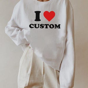 Op de afbeelding: Witte trui met een zwarte afbeelding die "I &hearts; CUSTOM" zegt. Het hart is rood.