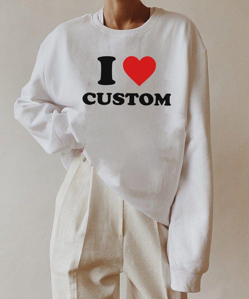 Op de afbeelding: Witte trui met een zwarte afbeelding die "I &hearts; CUSTOM" zegt. Het hart is rood.