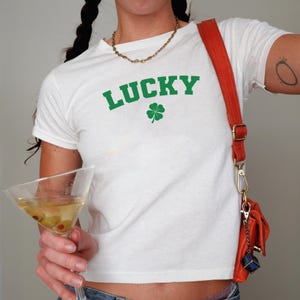 Può includere: T-shirt bianco con la scritta "LUCKY" in lettere verdi e un quadrifoglio verde. La maglietta è abbinata a una borsa a tracolla rossa e una collana a catena dorata. Un bicchiere da martini con olive è tenuto in mano.