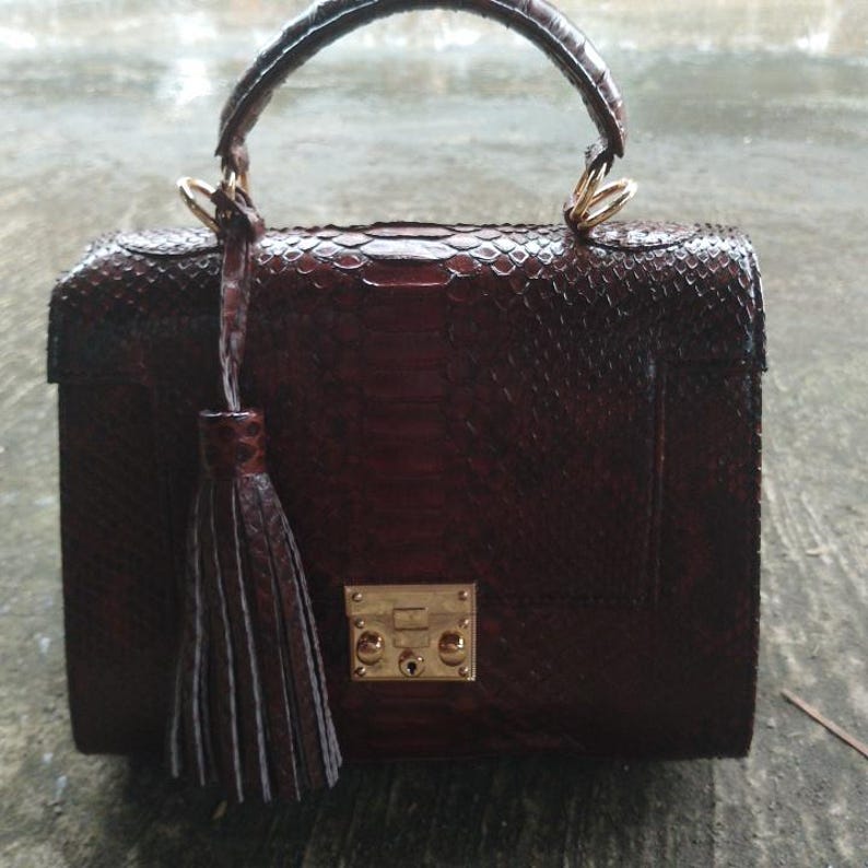 Genuine clutch python leather top-handle handbag featuring polished gold-tone hardware and a matching leather tassel, displayed against a rustic bamboo and wood background