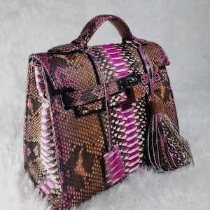 May include: A handcrafted handbag with a snakeskin pattern in shades of brown, pink, and white. The bag features a top handle, a flap closure with a silver-tone clasp, and a decorative tassel. The bag is a stylish accessory.