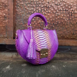 Op de afbeelding: Een levendige paarse handtas met een afgeronde vorm en een textuur die op slangenleer lijkt. De tas heeft een handvat aan de bovenkant, een afneembare schouderriem en een decoratieve kwast. Goudkleurige hardware accentueert de sluiting.