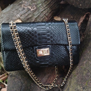 Könnte beinhalten: Eine schwarze Ledertasche mit einer goldenen Kettenriemen und einem goldenen Verschluss. Die Tasche hat ein Schlangenhautmuster.