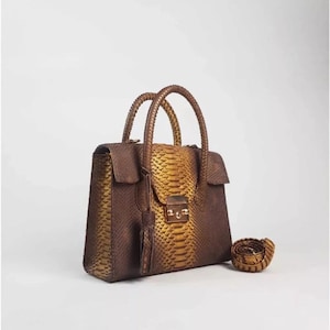 Könnte beinhalten: Eine braune und goldene Schlangenleder-Handtasche mit einem Henkel oben und einem abnehmbaren Schulterriemen. Die Tasche hat ein goldenes Schloss und einen Quasten.
