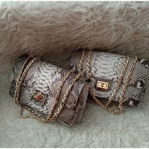 Może przedstawiać: Dwie torebki ze wzorem skóry węża w odcieniach szarości i brązu. Każda torba posiada złoty łańcuszek i prostokątne zapięcie. Torebki są prezentowane na puszystej, białej powierzchni.