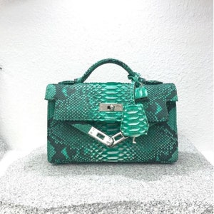 Könnte beinhalten: Eine grüne Handtasche aus Schlangenhaut mit einem oberen Griff und einem Klappenverschluss. Die Tasche hat silberfarbene Hardware und einen passenden Schlangenhaut-Anhänger. Die Tasche steht auf einer grauen Oberfläche.