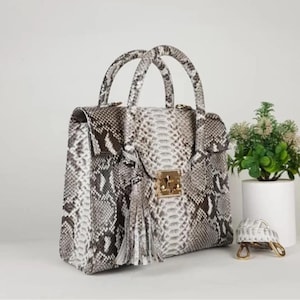 以下が含まれることがあります： グレー、白、茶色のスネークスキン柄の構造化されたハンドバッグ。バッグはトップハンドル、ゴールドカラーの留め金付きのフラップクロージャー、装飾的なタッセルが特徴です。バッグの隣には、小さな装飾的なカメの置物があります。