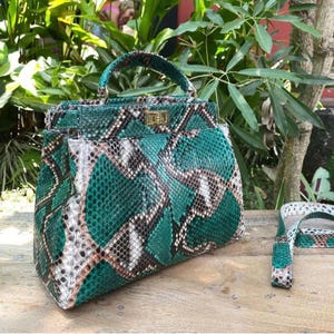 May include: A green, brown, and white snakeskin-patterned handbag with a top handle and a detachable shoulder strap. The bag features a gold-colored clasp and hardware. The bag is sitting on a wooden surface.