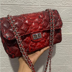 Può includere: Una borsa rossa trapuntata con tracolla e chiusura a catena argentata. La borsa è realizzata in un materiale simile alla pelle di serpente. La borsa è tenuta da una mano.