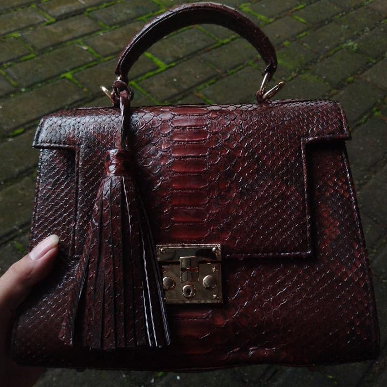 Genuine 100% python leather top-handle handbag featuring polished gold-tone hardware and a matching leather tassel, displayed against a rustic bamboo and wood background