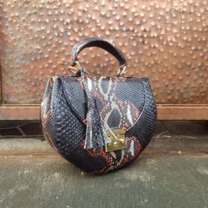 Puede incluir: Un bolso de mano negro y rojo con estampado de piel de serpiente, de forma redondeada y con asa superior. El bolso presenta un cierre dorado y una borla. El bolso está hecho de un material texturizado.