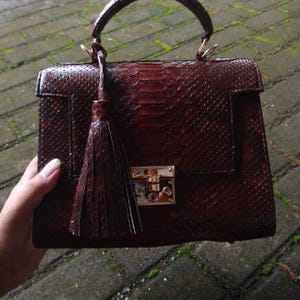 Genuine small python leather top-handle handbag featuring polished gold-tone hardware and a matching leather tassel, displayed against a rustic bamboo and wood background