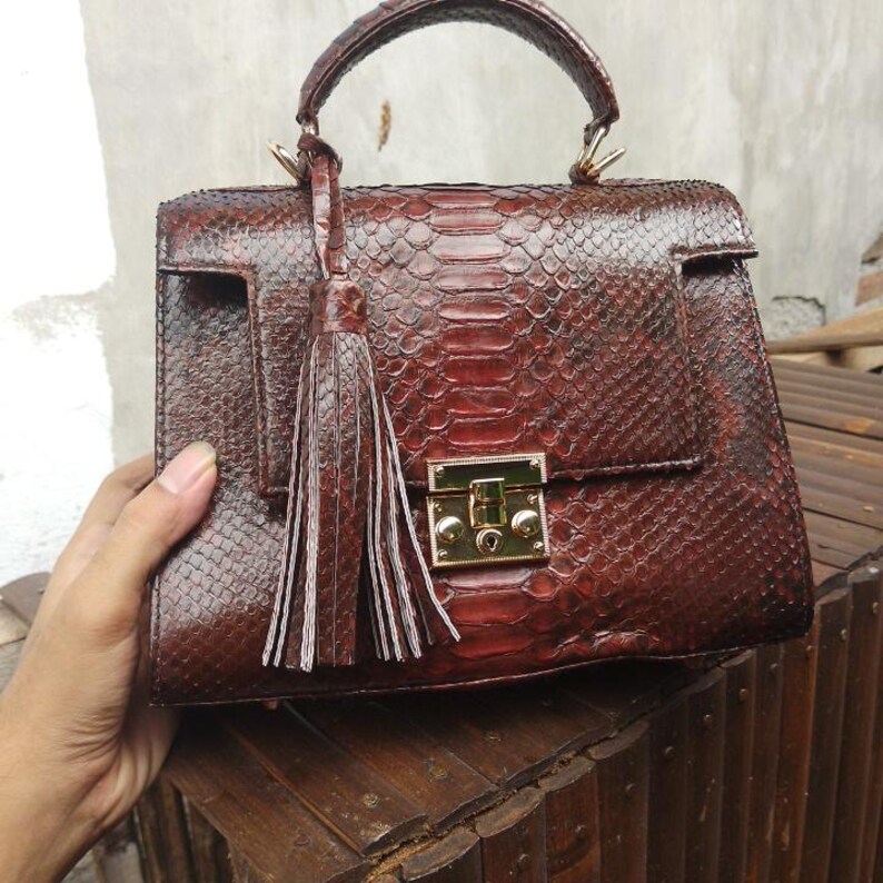 Genuine adorable python leather top-handle handbag featuring polished gold-tone hardware and a matching leather tassel, displayed against a rustic bamboo and wood background