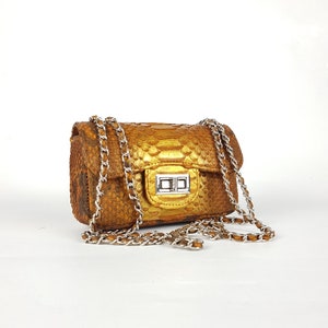 Peut inclure: Petit sac à main rectangulaire avec une bandoulière chaîne. Le sac est fait d'un matériau texturé brun doré avec un fermoir argenté. La bandoulière chaîne est argentée. Le sac est sur fond blanc.