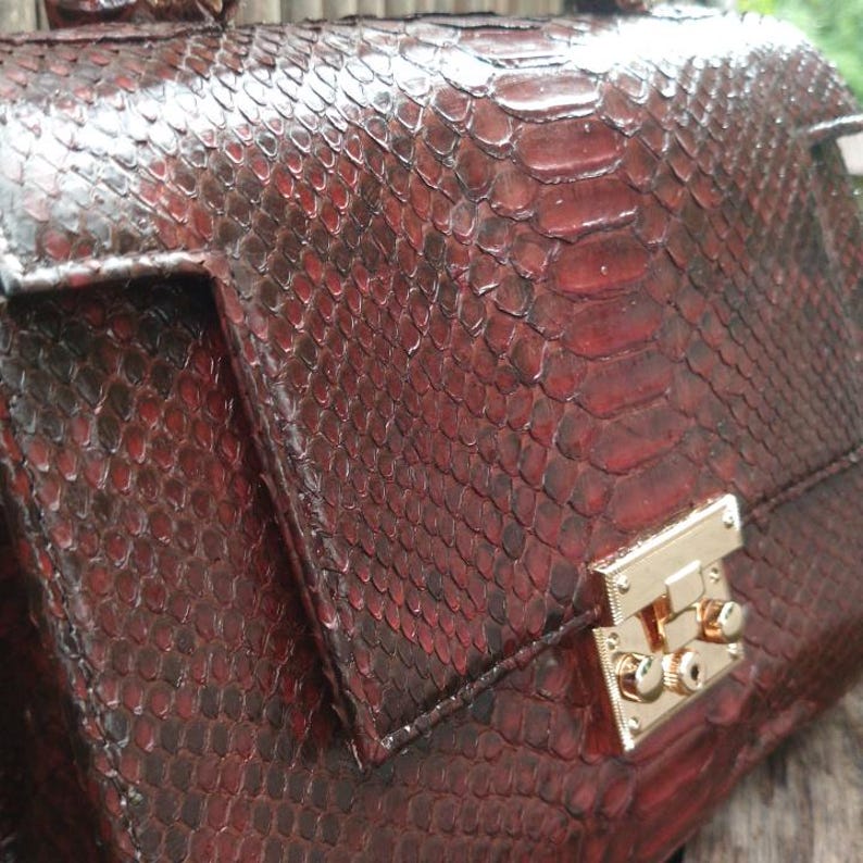 Genuine maroon red python leather top-handle handbag featuring polished gold-tone hardware and a matching leather tassel, displayed against a rustic bamboo and wood background
