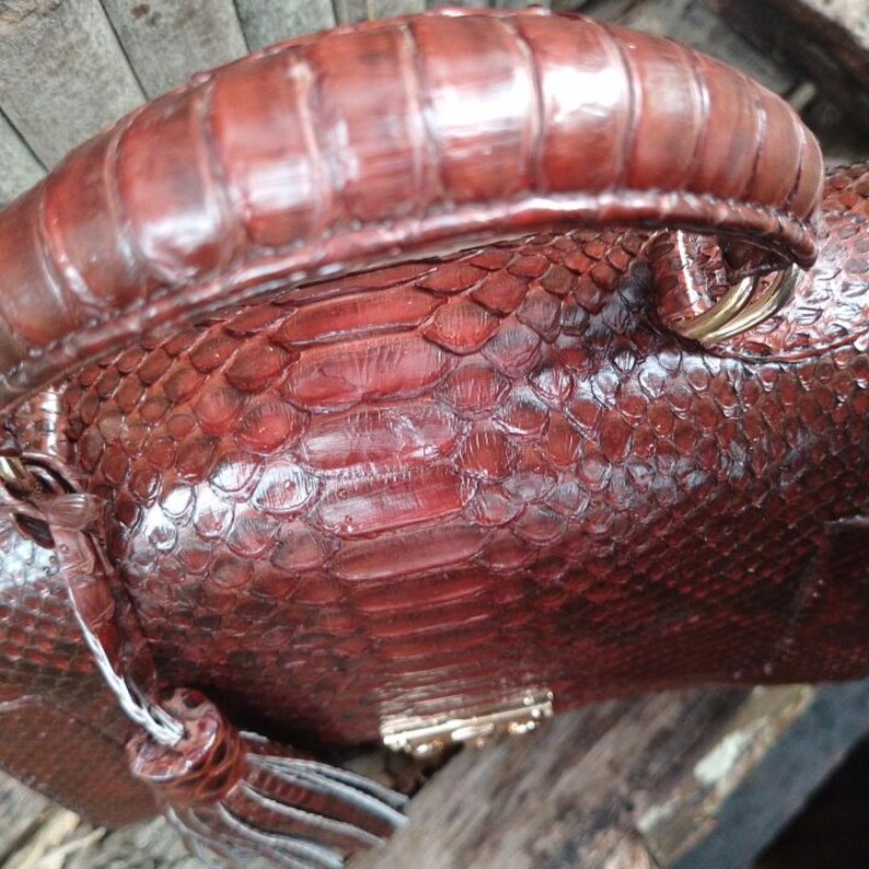 Genuine maroon python leather top-handle handbag featuring polished gold-tone hardware and a matching leather tassel, displayed against a rustic bamboo and wood background